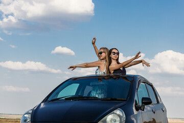 Beautiful young women in sunglasses dance against the background of a blue sky with clouds leaning out of the car hatch. The concept of traveling by private car