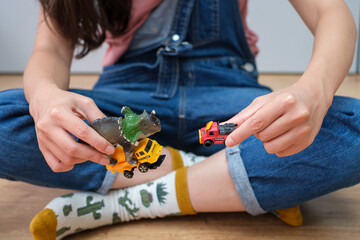 Girl on wooden floor playing with cars and dinosaurs toys.