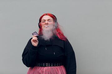 European plus size woman in headphones smoking vape electronic cigarette. Young red pink haired...