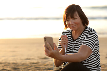 Naklejka premium Smiling mature caucasian woman listening music in headphones looking at camera relaxing and resting on sandy beach by the sea at sunrise. Relaxation concept