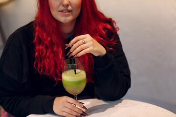 European plus size woman drink green smoothie in cafe. Young red pink haired body positive girl...