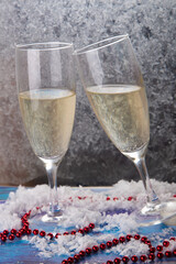 Merry Christmas, happy new year greeting card. Two nice tall wineglasses with white wine stay on frozen window background. Snow, red beads on wooden blue table. Vertical photo, selective focus