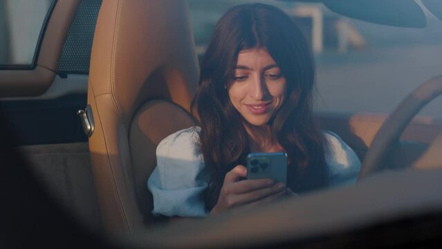 Beautiful stylish woman sits in her luxury white convertible and uses smartphone for chatting. View through the windshield. Sunset.
