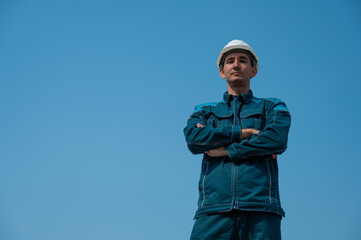 A caucasian man in work clothes and a construction helmet stands against the blue sky with his arms crossed on his chest. 