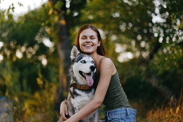 Joyful woman with a husky breed dog smiling while sitting in nature on a walk with a dog on a leash...