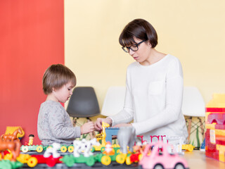 Obraz na płótnie Canvas Toddler plays with colorful toy blocks while his mother or babysitter. Little boy stares on toy constructor. Interior of kindergarten or nursery.