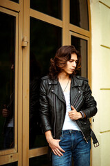 a young, handsome man with long dark hair, wearing a leather jacket, jeans and a white T-shirt, walks alone in the city