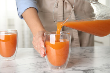 Woman pouring freshly made carrot juice into glass on white marble table in kitchen, closeup
