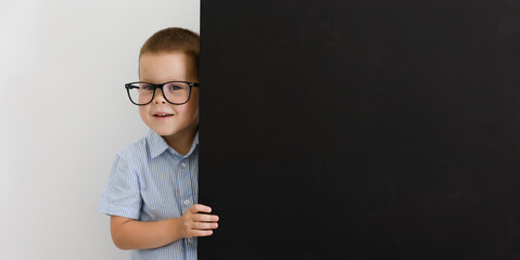 A child in glasses and a blue shirt peeks out from behind a slate. 