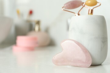 Rose quartz gua sha tool near holder with natural face rollers on white countertop in bathroom,...