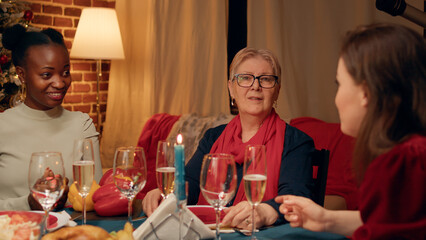 Happy senior woman sitting at Christmas dinner table with family members while looking at camera. Festive elderly person celebrating winter traditional holiday with close relatives at home.