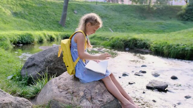 a little girl draws with pencils in an album sitting on a large stone near the river