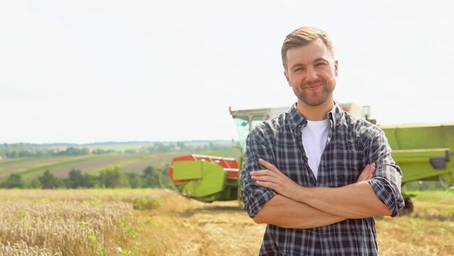 Happy young farmer standing on wheat field while combine harvester in background
