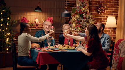 Cheerful multiethnic group of people celebrating winter holiday with sparkling wine. Happy close family members clinking champagne glasses while sitting together at Christmas dinner table. Tripod shot