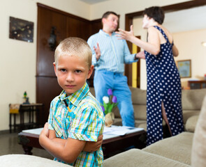 Obraz premium Unhappy small boy sitting at sofa during his parents quarrelling on background at home