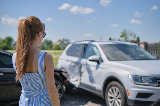 Sad young woman driver standing near her smashed car looking shocked on crashed vehicles in road accident