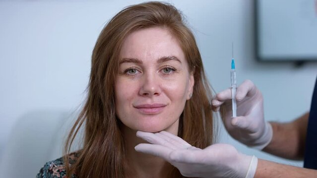 Front view headshot of confident woman looking at camera with plastic surgeon hand touching chin holding syringe. Beautiful Caucasian female patient posing indoors before beauty injection