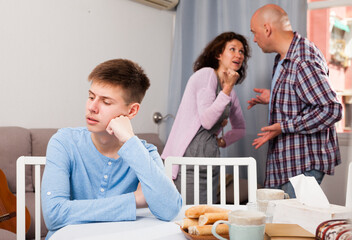 Portrait of upset teenage boy sitting at table while parents quarreling at home