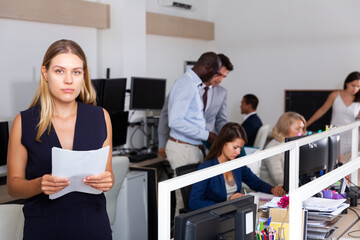 Puzzled and confused woman standing with papers in hands in open plan office on background with busy colleagues