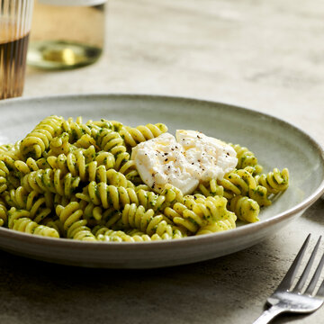 Bowl of pesto pasta topped with creamy burrata next to a glass o