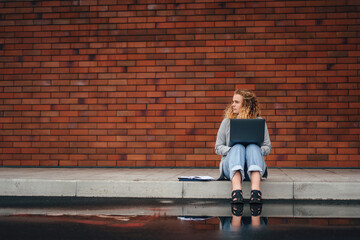 Fototapeta na wymiar Caucasian girl having training course via notebook device, sitting outdoors in city street on the background of a brick wall with free space for text