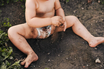 Baby girl only in a diaper got dirty while playing in the ground, exploring the world around. Happy family outdoors. Child care. Body care. - obrazy, fototapety, plakaty