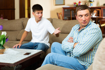 Portrait of worried father at odds with teenager son sitting on sofa at home