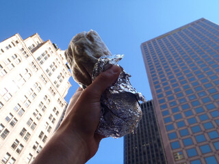 Hand holds Burrito wrapped in tinfoil in the air in downtown of city