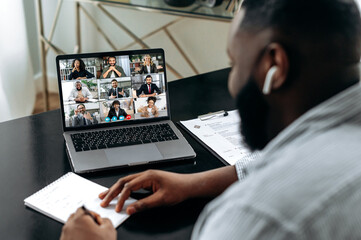 Friendly african american man, sit at a desk in, having video meeting with colleagues, conducts financial brainstorm with business partners, takes notes. Group video meeting with multiracial people