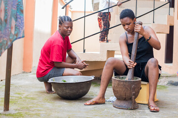 African females, ladies or women from Nigeria doing house chores while one is using a wooden pestle and mortar to pound and grind edible vegetables for food cooking, herb or medications - obrazy, fototapety, plakaty
