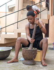 An African young girl, lady or woman from Nigeria using a wooden pestle and mortar to pound and grind edible vegetables for food cooking, herb or medications