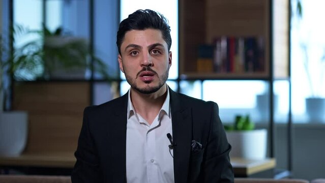 Elegant successful man talking looking at camera sitting in comfortable modern office. Front view portrait of confident intelligent Middle Eastern businessman posing giving interview indoors