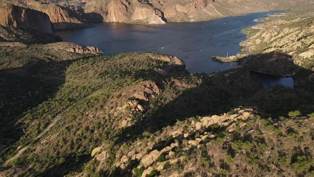 Drone flying over mountains and then slowly panning up revealing a dark blue lake with boats on it, late evening, golden hour, apache lake