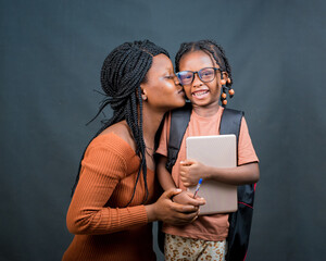 An african Nigerian mother, teacher or guardian, kissing a girl child or daughter on the cheek while the happy little one is holding an education smart tablet and also carrying a school back pack