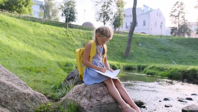 a girl draws in an album sitting on a large stone on the river bank
