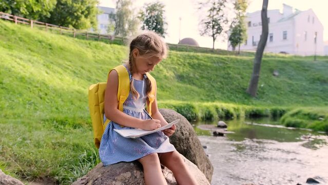 a girl draws a picture sitting on a large stone on the bank of a fast river