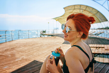 Red-haired girl in sunglasses smiles sweetly and rejoices. Woman with bottle of juice and straw is sitting on chaise longue on pier. Blue sea around. Sunny weather. Copy space for travel advertising.