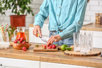 Woman cutting strawberry for tasty lemonade at table in kitchen