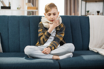 Cold and flu season. Sick teenage boy with high fever and sore throat and feeling bad, sitting at home on the sofa. Little child touches his throat with his hand feeling pain. - obrazy, fototapety, plakaty