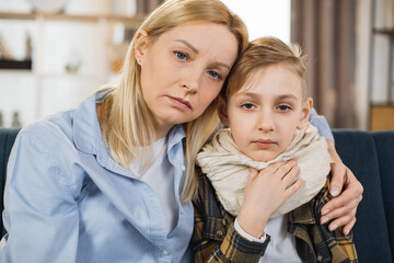 Close up portrait of sad sick teen boy with scarf around neck and his worried young mother, sitting...
