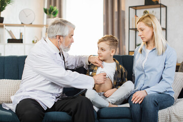 Preschool sick cute boy sitting with his mother on the couch while caring experienced nice mature male doctor listening his heartbeat during home visit, front view.