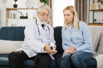 Fototapeta premium Caucasian male senior doctor using infrared thermometer gun, checking the body temperature of his sick woman patient, while visiting her at home.