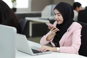 Attractive professional asian muslim female employee worker sitting, using laptop computer with coworker at workplace. Businesswoman use speaker phone on smart mobile phone and communicate with client