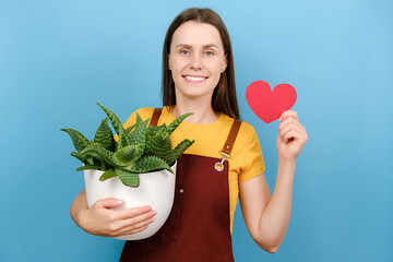Attractive young female florist holding red heart and pot plant, happy looking at camera, posing...