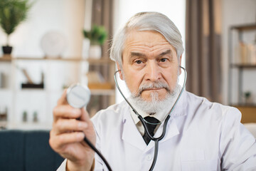 Qualified senior doctor in eyewear and lab coat holding stethoscope while posing at conference room. Concept of people, medicine and occupation.