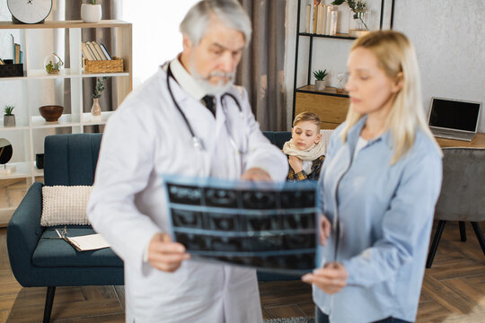 Professional mature man doctor explaining results of CT x-ray image of teen boy to his worried mother, while visiting patient at home. Focus on sick child sitting on sofa on the background.