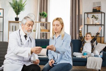 Pretty caring mother sitting with experienced confident mature bearded male doctor and listening instructions about use of medications, while excited boy jumping on the bed rejoicing at the recovery