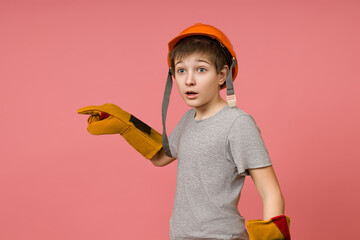 shocked child in a hard hat and construction gloves points a finger to the side on a pink background with copy space