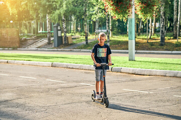 Schoolboy rides electric scooter on road in empty summer park. Child in black t-shirt and denim shorts rides on scooter along marked asphalt road
