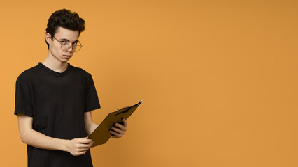 serious young man in glasses holding a paper clipboard on a yellow background with copy space